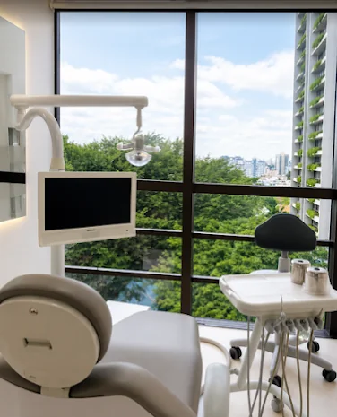 Sala de atendimento com cadeira odontológica, monitor e janela do chão ao teto com vista para árvores e prédios
